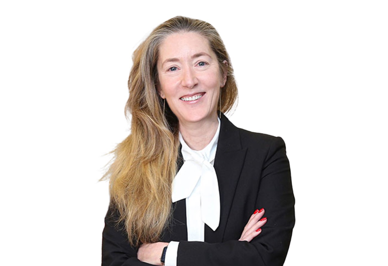 Smiling woman with long hair, wearing a black blazer and white blouse, standing with arms crossed against a white background.